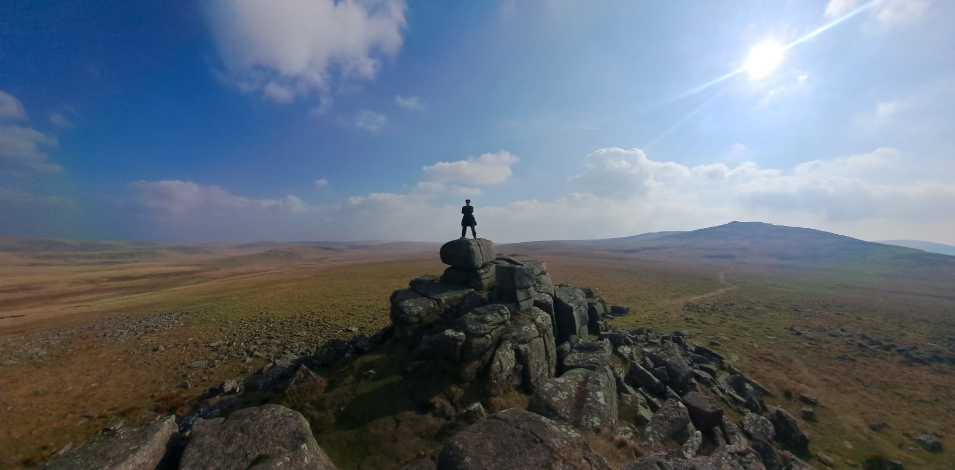 A figure standing on a granite tor on Dartmoor, with sweeping moorland under a bright sky